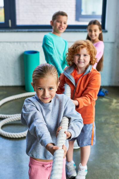 four cheerful preadolescent kids playing tug of war and looking at camera in gym, child sport