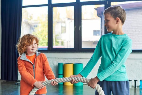two preadolescent boys in sportswear pulling fitness rope and smiling at each other, child sport