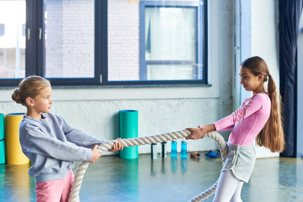 two pretty girls in sportswear pulling fitness rope in gym and looking at each other, child sport