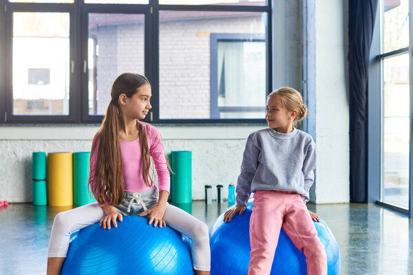 two pretty girls in sportswear exercising on fitness balls and looking at each other, child sport