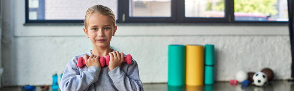 preadolescent cute blonde girl exercising with dumbbells and looking at camera, child sport, banner