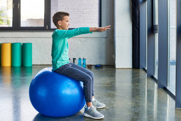 cute preadolescent boy in blue sportswear stretching and training on fitness ball, child sport