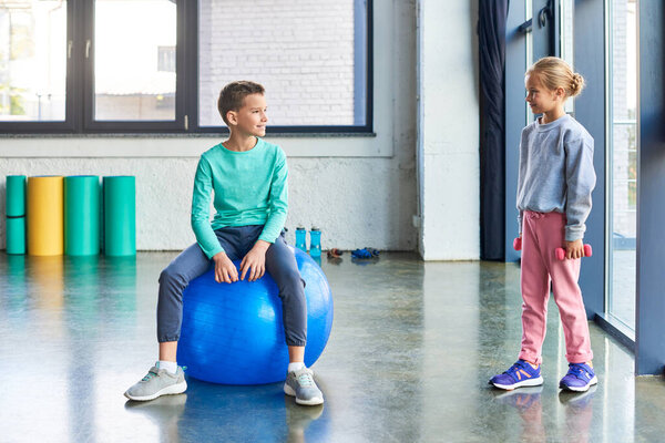 little blonde girl with dumbbells in hands smiling at cute boy on fitness ball, child sport
