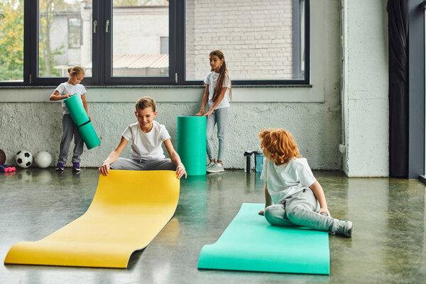 cheerful preadolescent children in sportswear unfolding fitness mats in gym, workout, child sport