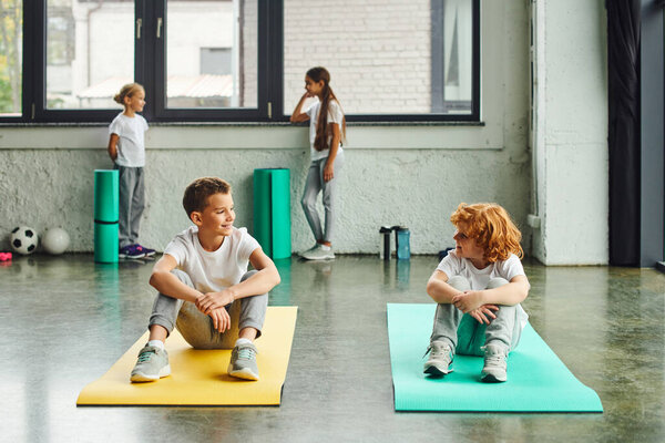 two cheerful boys smiling at each other on fitness mats with cute girls standing on backdrop