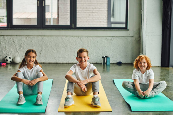 three cheerful preadolescent boys and girl sitting on fitness mats and smiling at camera, sport
