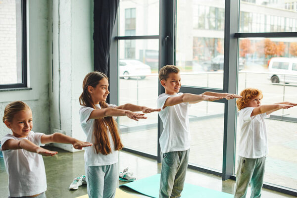 joyous little children in sportswear stretching their arms in gym with window backdrop, child sport
