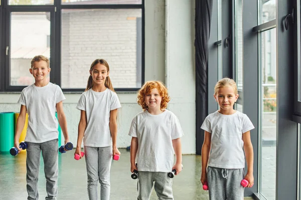 Stock image four cheerful little children posing with dumbbells and smiling joyfully at camera, child sport