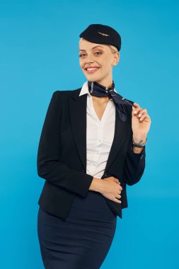 happy and pretty air hostess in uniform touching elegant neckerchief and looking at camera on blue clipart