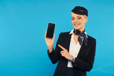 elegant air hostess in stylish uniform pointing at smartphone with blank screen on cyan backdrop clipart