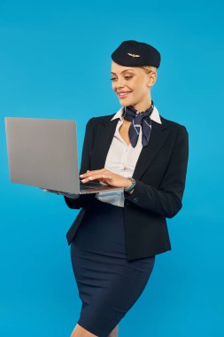 Mavi arka planda dikilirken dizüstü bilgisayar kullanan zarif hostes üniformalı neşeli genç bir kadın.