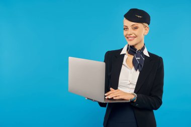 Şık şirket üniformalı neşeli hostes elinde dizüstü bilgisayarla mavi arka planda kameraya bakıyor.