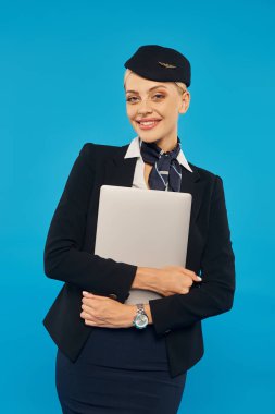 charming flight attendant with radiant smile holding laptop and looking at camera on blue background clipart