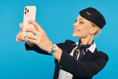 Uçuş görevlisi üniformalı mutlu genç bayan mavi arka planda cep telefonuyla selfie çekiyor.