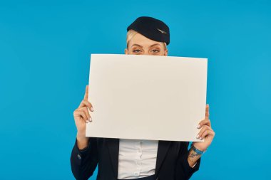 Hostes üniformalı genç bir kadın. Mavi arka planda boş bir afişle yüzünü gizliyor.