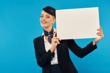 Şık üniformalı neşeli hostes elinde boş levhayla mavi arka planda kameraya gülümsüyor.