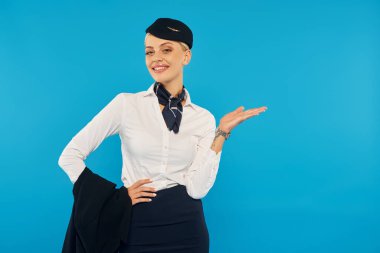 Beyaz bluzlu mutlu hostes ve hostes şapkası elinle gösterip mavi ceketini tutuyor.