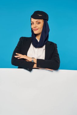 Arap havayolları hostesi kollarını kavuşturmuş mavi arka plandaki boş posterin yanında kameraya gülümsüyor.
