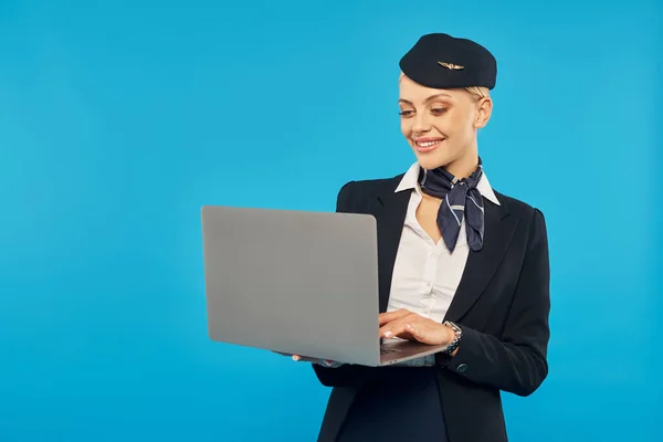 young and smiling air hostess in stylish uniform networking on laptop on blue studio backdrop