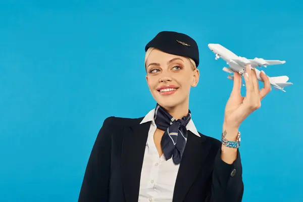 joyous air hostess in uniform of commercial airlines posing with airplane model on blue backdrop