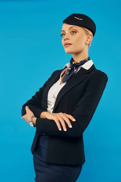 confident woman in elegant uniform of air hostess posing with folded arms and looking away on blue