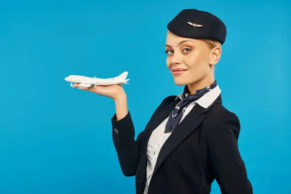 young smiling woman in stewardess uniform holding airplane model and looking at camera on blue