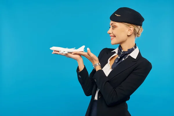 cheerful attractive stewardess in uniform standing with airplane model on blue backdrop, side view