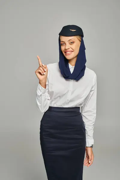young arabian airlines stewardess in headscarf and uniform pointing up with finger on grey backdrop