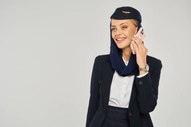 Arap havayollarında genç ve zarif hostes gri zeminde cep telefonuyla konuşuyor.