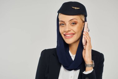 Arap havayolları üniformalı çekici hostes akıllı telefondan konuşuyor ve gri kameraya gülümsüyor.