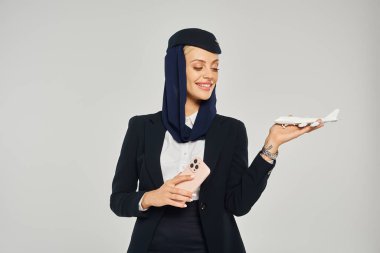 Gülümseyen Arap havayolları hostesi cep telefonuyla gri zeminde uçak modeline bakıyor.