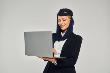 memnun Arap havayolları hostesi şirket üniforması içinde dizüstü bilgisayar kullanarak gri üzerinde dururken