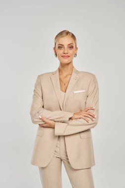 Bej elbiseli gülümseyen kadın kollarını kavuşturup gri renkli moda modasına bakıyor.