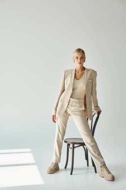 Modayı takip eden sarışın kadın kameraya bakarken gri arka planda, güneş ışığı altında sandalyeyle poz veriyor.