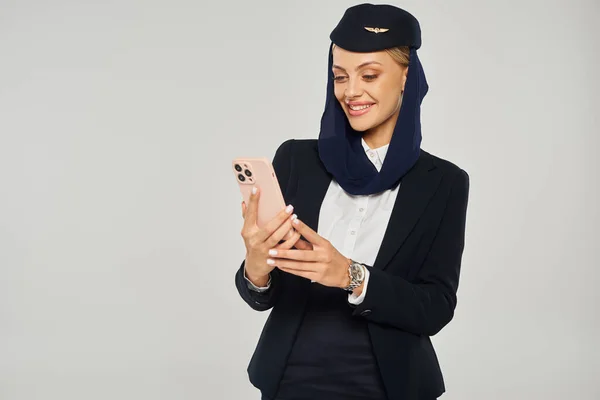 stock image smiling stewardess in uniform of arabian airlines messaging on mobile phone on grey backdrop