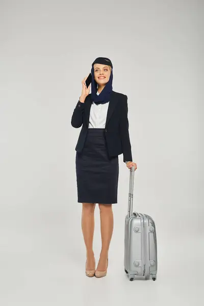 Stock image pleased arabian airlines air hostess with travel bag talking on mobile phone on grey backdrop