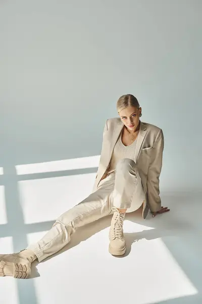 stock image charming woman in total beige outfit sitting in sunlight and looking at camera on grey, full length