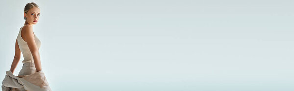 sensual blonde woman in beige tank top holding blazer and looking at camera on grey, banner