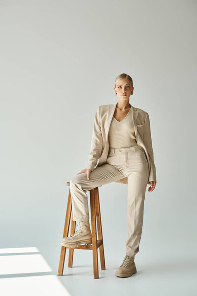 young elegant woman looking at camera while posing with tall stool on grey backdrop with sunlight