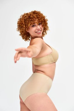 cheerful and redhead woman in taupe lingerie pointing and looking at camera on white, curvy body clipart