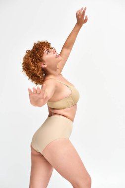 happy plus size redhead woman in beige lingerie standing and looking up in expressive pose on white clipart