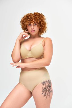 sensual tattooed plus size woman in taupe underwear with red wavy hair looking at camera on white clipart