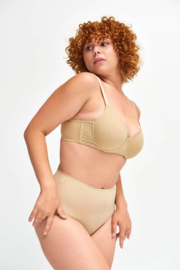 body positive redhead and curly woman in taupe underwear looking away on white, self-expression clipart