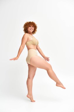 full length of happy plus size model in beige underwear standing and looking at camera on white clipart