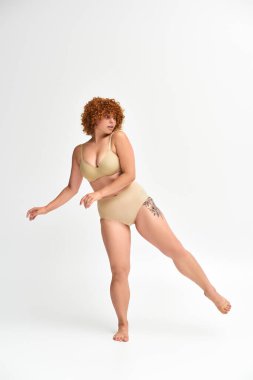 young and curvy model with tattooed hip posing in taupe lingerie on white backdrop, full length clipart