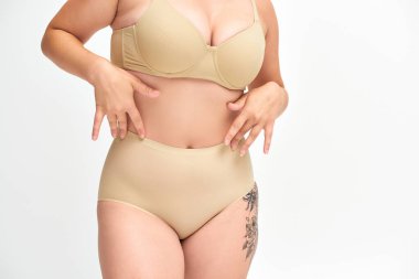 cropped view of young plus size woman in beige lingerie with tattooed hip on white backdrop clipart