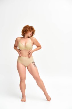 full length of alluring redhead woman with curvy tattooed body on white, non-traditional beauty clipart