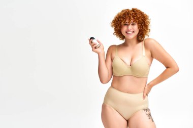 Bej iç çamaşırlı neşeli kadın deodorant spreyi tutuyor ve beyaz kameraya gülümsüyor.