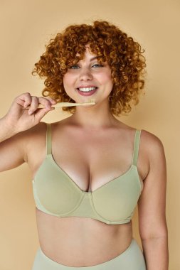 cheerful redhead woman with curvy body holding toothbrush and smiling at camera on beige backdrop clipart