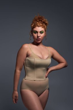 Çekici kızıl saçlı, bej renkli iç çamaşırlı, elleri koyu gri arka planda poz veren bir kadın.